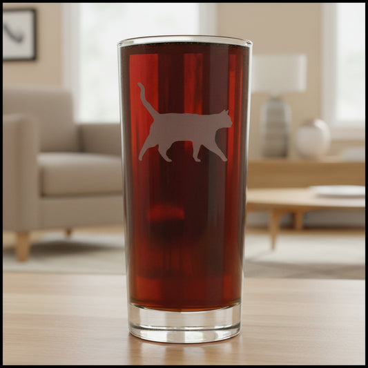 A clear pint glass filled with dark beer, featuring a etched cat silhouette on the front, set against a living room background.