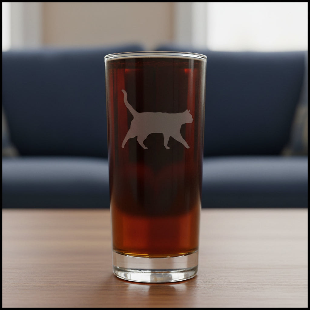 A clear pint glass filled with dark beer, featuring an etched cat silhouette on the front, set against a tblue couch.