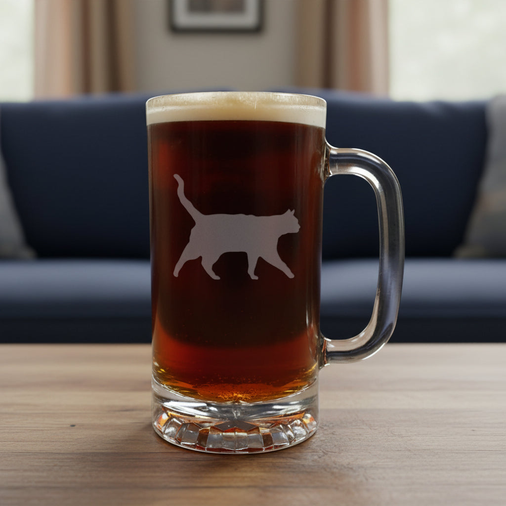 A clear glass beer mug filled with dark beer and topped with white foam, featuring a cat silhouette etched on the front, set on a light wood surface against a blue couch background.