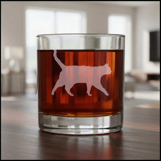 A clear rocks glass filled with an amber-red liquid, featuring a etched  cat silhouette design on the front, set against a modern living room.