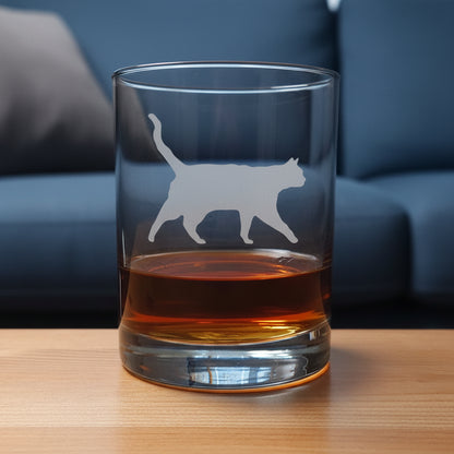 A clear glass rocks tumbler with a cat silhouette etched on the front, partially filled with amber whiskey, set on a light wood surface against a blue couch.