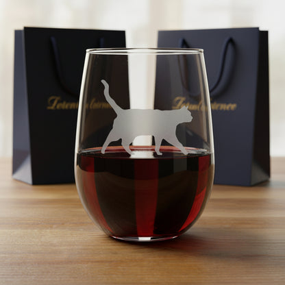 A clear stemless wine glass with a cat silhouette etched on the front, shown standing on a wooden surface with two gift bags in the background.
