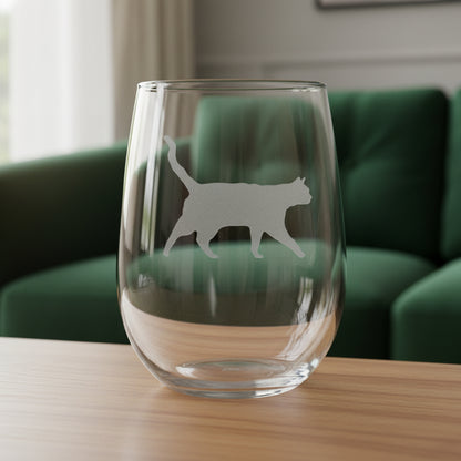 A clear stemless wine glass with a cat silhouette etched on the front, shown standing on a wooden surface against a green couch background.