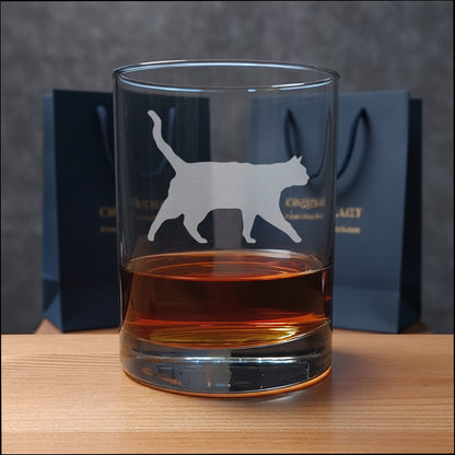 A clear glass rocks tumbler with a cat silhouette etched on the front, partially filled with amber whiskey, set on a light wood surface with two gift bags in the background.