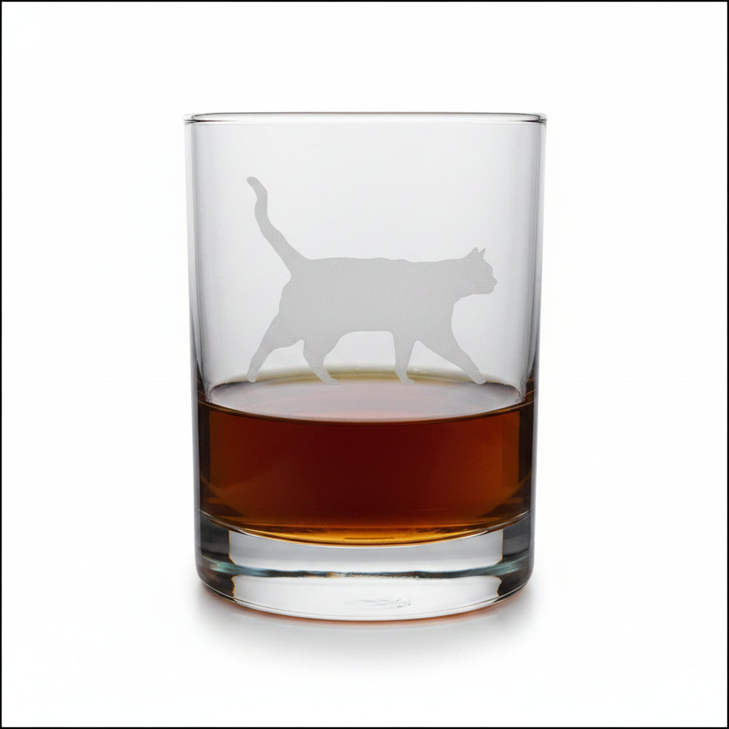 A clear glass rocks tumbler with a cat silhouette etched on the front, partially filled with amber whiskey, set on a light wood surface against a white background.