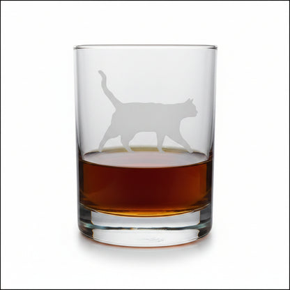 A clear glass rocks tumbler with a cat silhouette etched on the front, partially filled with amber whiskey, set on a light wood surface against a white background.