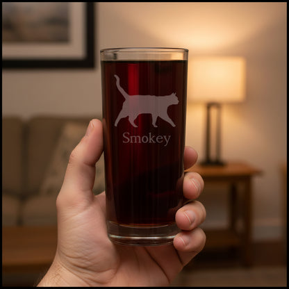A hand holds a clear pint glass filled with dark beer, featuring an etched silhouette of a cat and the name 'Smokey' on the front, set against aliving room background.