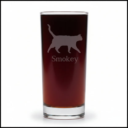 A clear pint glass filled with dark beer, featuring an etched silhouette of a cat and the name 'Smokey' on the front, set against a white background.