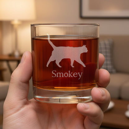 A hand holding a clear glass rocks tumbler filled with amber-red liquid, featuring an etched silhouette of a walking cat and the name 'Smokey' on the front, set against a living room.