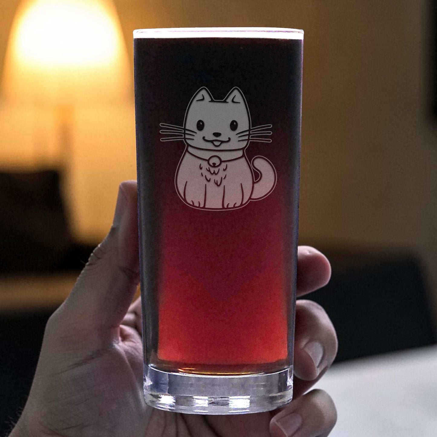 A hand holds a clear glass cooler glass filled with a dark red beverage. The glass features an etched cute cat wearing a collar.