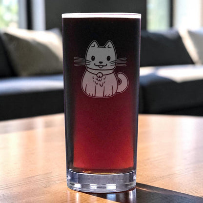 A tall, clear pint glass with a cute cat illustration on the front, filled with a dark reddish-brown beverage and set on a wooden table in a softly blurred living room setting.