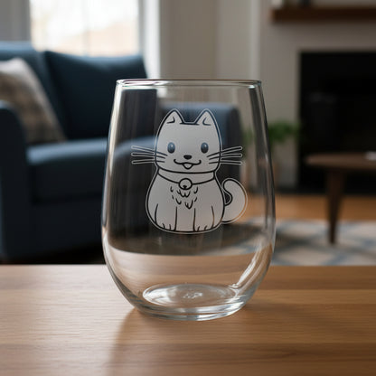 A clear stemless wine glass with an etched cute cat design on the front, set on a wooden surface with a blue couch in the background.