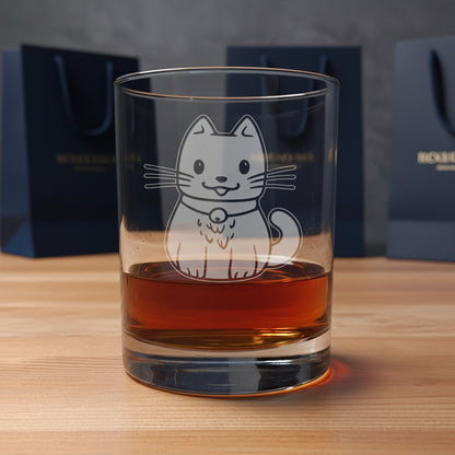 A clear glass rocks tumbler with an etched design  of a cute cat wearing a bell, shown filled with amber whiskey on a wooden surface with gift bags in the background background.