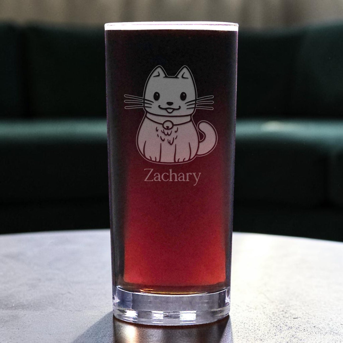 A clear pint glass filled with a dark red drink, featuring an etched cute cat and the name 'Zachary' on the front, set on a tabletop with a blurred dark green couch in the background.