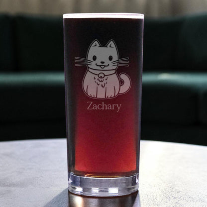 A clear pint glass filled with a dark red drink, featuring an etched cute cat and the name 'Zachary' on the front, set on a tabletop with a blurred dark green couch in the background.