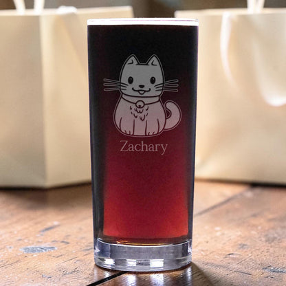 A clear glass tumbler with a red gradient insert, featuring a cute cat and the name 'Zachary' printed on the front. The glass is set on a rustic wooden table with blurred cream-colored gift bags in the background.