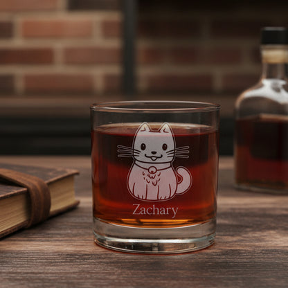 A clear rocks glass filled with a dark red drink, featuring an etched  cat design and the name 'Zachary' on the front, set against a brick  background with a bottle of liquor to the right and a book to the left.