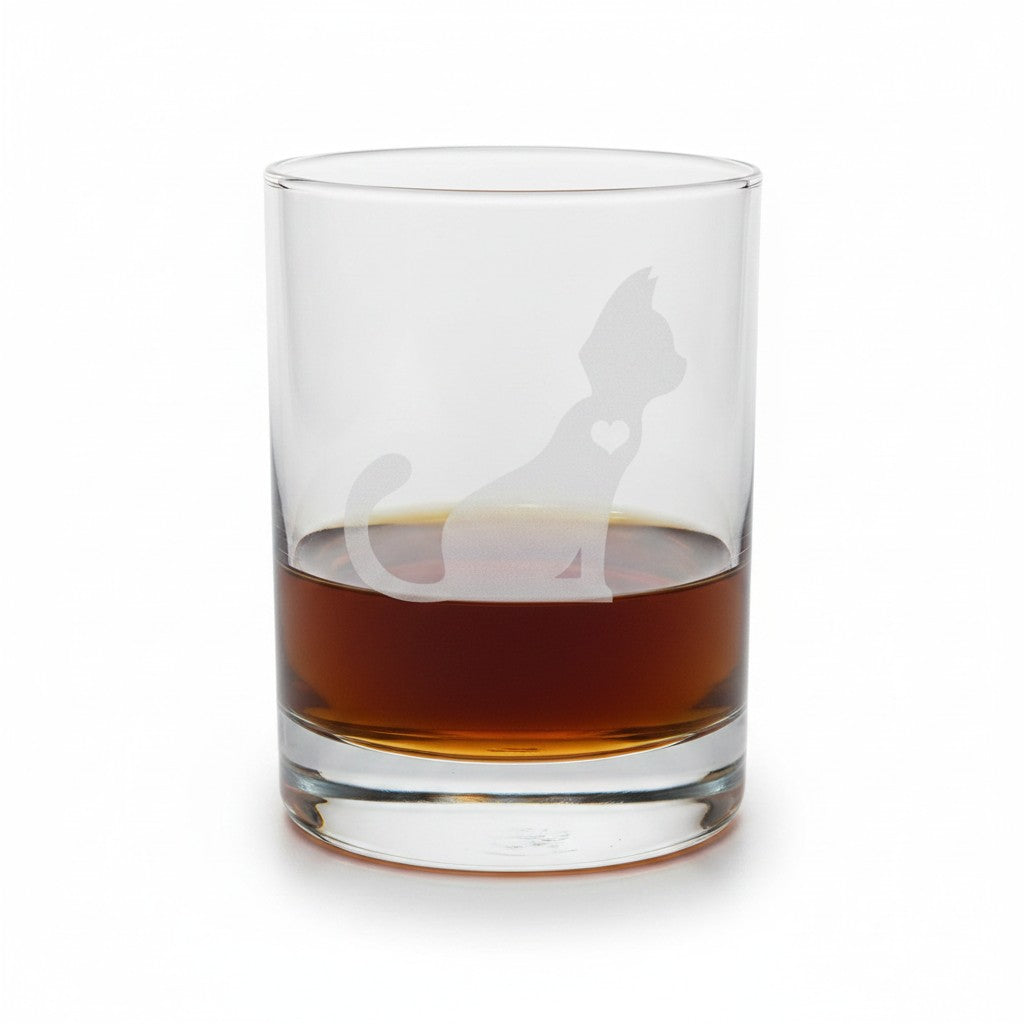 A clear glass rocks tumbler with an etched silhouette of a cat with heart, shown filled with an amber-brown liquid on a white background.