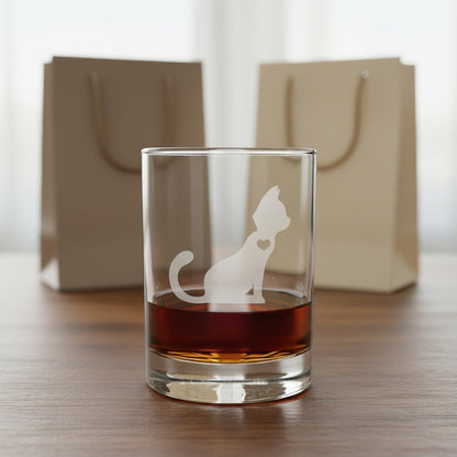 A clear lowball tumbler glass with an etched cat silhouette and heart design, partially filled with amber liquor, set on a wooden table with two beige gift bags blurred in the background.