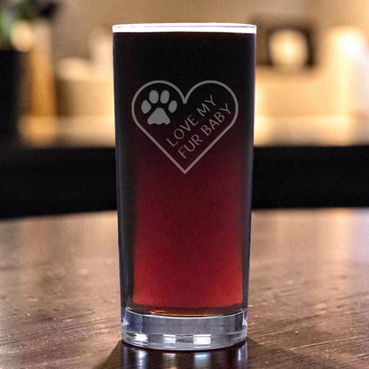 A clear pint glass filled with dark red beer, featuring a etched heart-shaped design with a paw print and the text 'LOVE MY FUR BABY', set on a wooden table with a blurred indoor background.