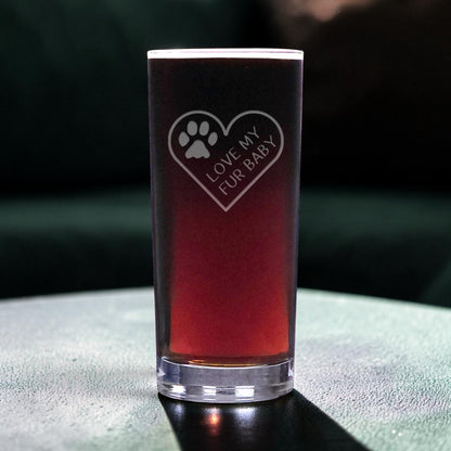 A clear highball glass filled with a dark reddish-brown liquid, featuring an etched heart design with a paw print and the words 'LOVE MY FUR BABY'. The glass is set on a dark, textured surface with a dark, moody background.