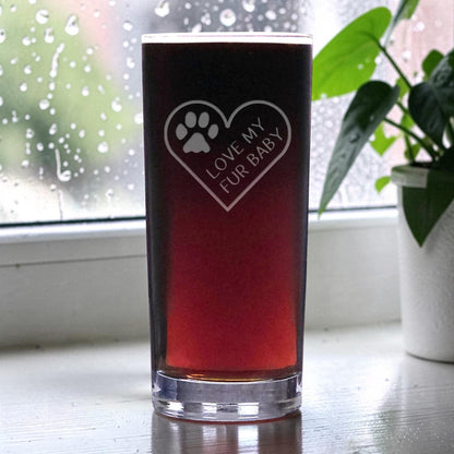 A clear pint glass filled with dark red beer, featuring a etched heart design with a paw print and the text 'LOVE MY FUR BABY', set on a windowsill with raindrops and a potted plant in the background.