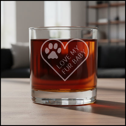 A clear glass rocks tumbler filled with amber liquid, featuring an etched heart design with a paw print and the words 'LOVE MY FUR BABY' on the front, set against a modern living room background.