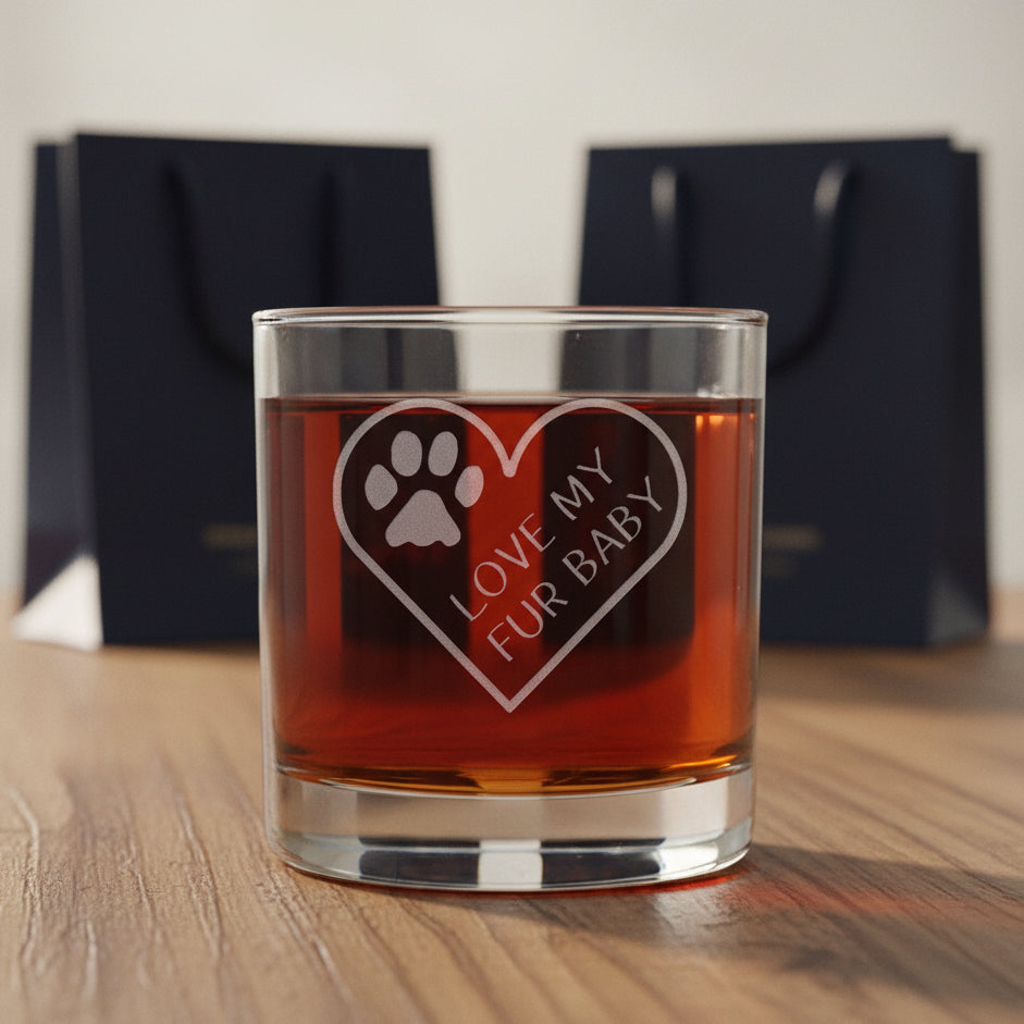 A clear glass rocks tumbler filled with amber liquid, featuring an etched heart design with a paw print and the words 'LOVE MY FUR BABY' on the front, two gift bags are in the  background.