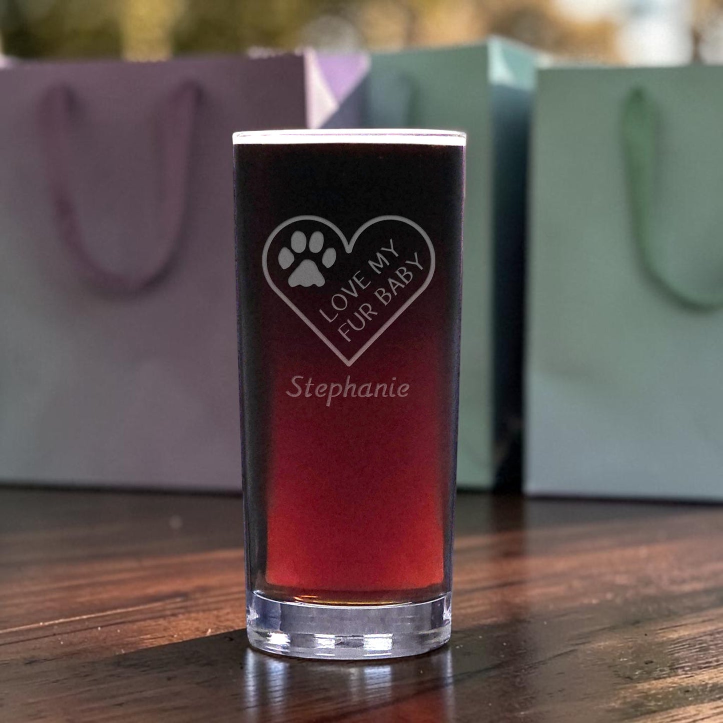 A clear glass shot glass filled with a dark red liquid, standing on a wooden table. The glass is etched with a heart, paw print, the phrase 'LOVE MY FUR BABY', and the name 'Stephanie'.