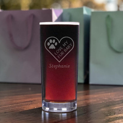 A clear glass shot glass filled with a dark red liquid, standing on a wooden table. The glass is etched with a heart, paw print, the phrase 'LOVE MY FUR BABY', and the name 'Stephanie'.