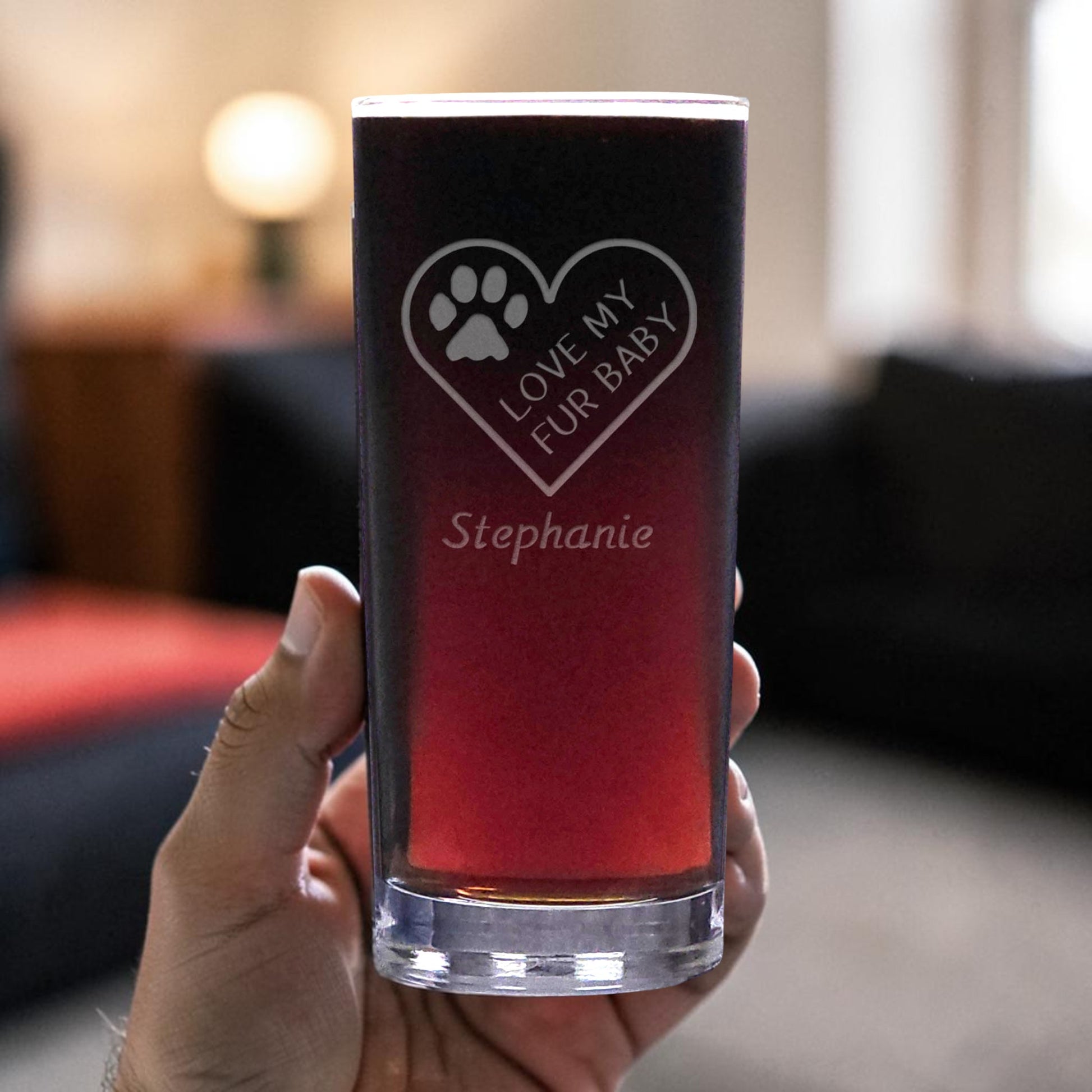 A hand holds a clear glass tumbler with a reddish liquid, featuring an etched  heart-shaped paw print and the text 'LOVE MY FUR BABY' and the name 'Stephanie'. The background is a softly blurred indoor living room setting.