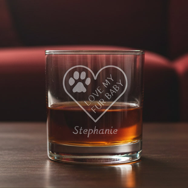 A clear glass rocks tumbler filled with amber liquid, featuring an etched heart design with a paw print and the words 'LOVE MY FUR BABY' along with the name 'Stephanie' below, set against a tred couch.