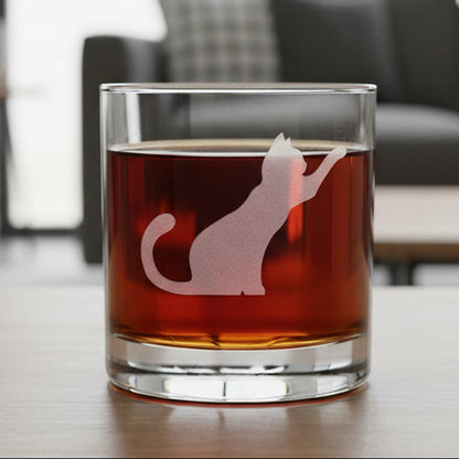 A clear rocks glass filled with dark red liquid, featuring an etched cat silhouette design on the front, set against a cozy living room