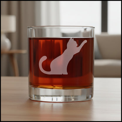 A clear rocks glass filled with dark red liquid, featuring a frosted white cat silhouette design on the front, with a living room in the background.