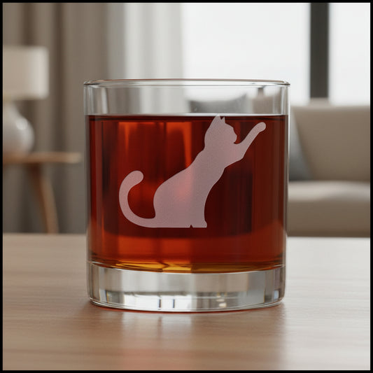 A clear rocks glass filled with dark red liquid, featuring a frosted white cat silhouette design on the front, with a living room in the background.