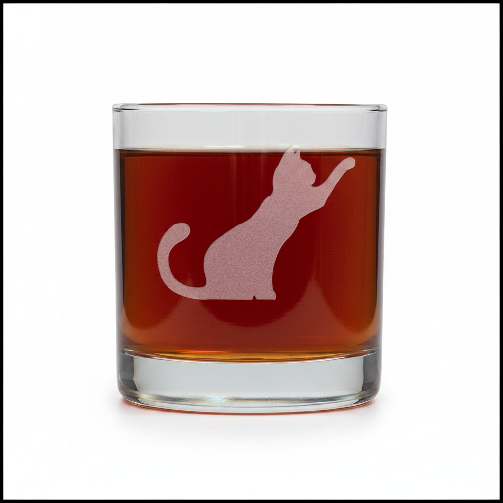 A clear rocks glass filled with dark red liquid, featuring a frosted white cat silhouette design on the front, set against awhite  background.