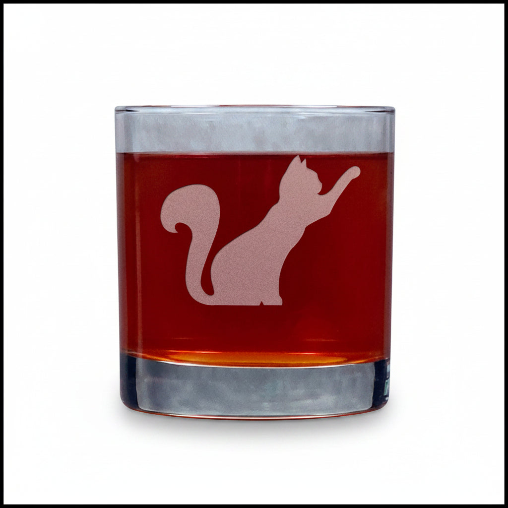 A clear rocks glass filled with amber-red liquid, featuring an etched silhouette of a stretching cat on the front, set against a textured gray background.