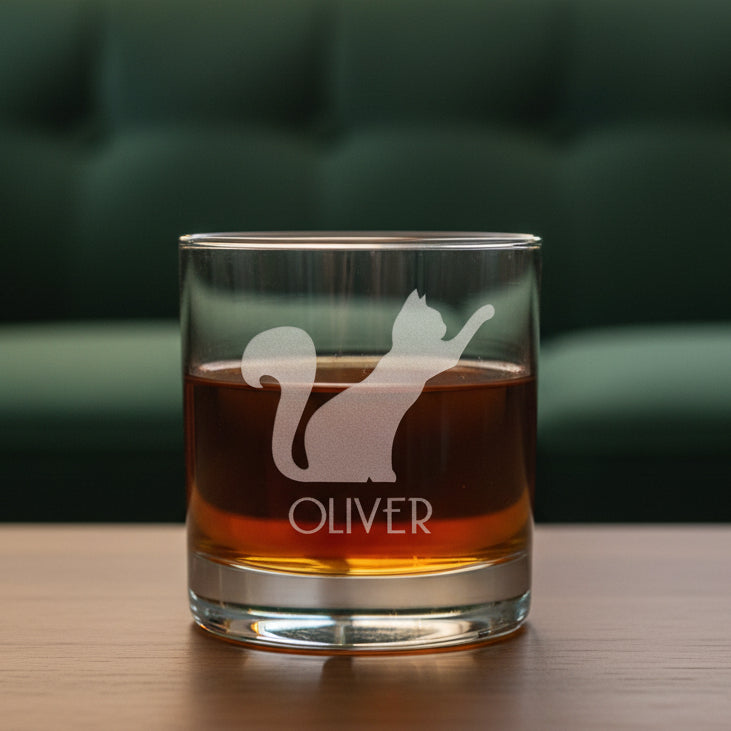 A clear glass rocks tumbler filled with amber liquid, featuring an etched  silhouette of a cat and the name OLIVER on the front, set against a green couch in the background.