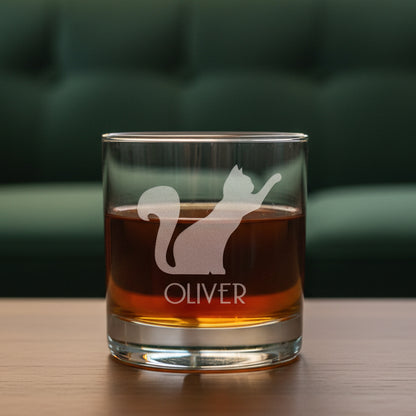 A clear glass rocks tumbler filled with amber liquid, featuring an etched  silhouette of a cat and the name OLIVER on the front, set against a green couch in the background.