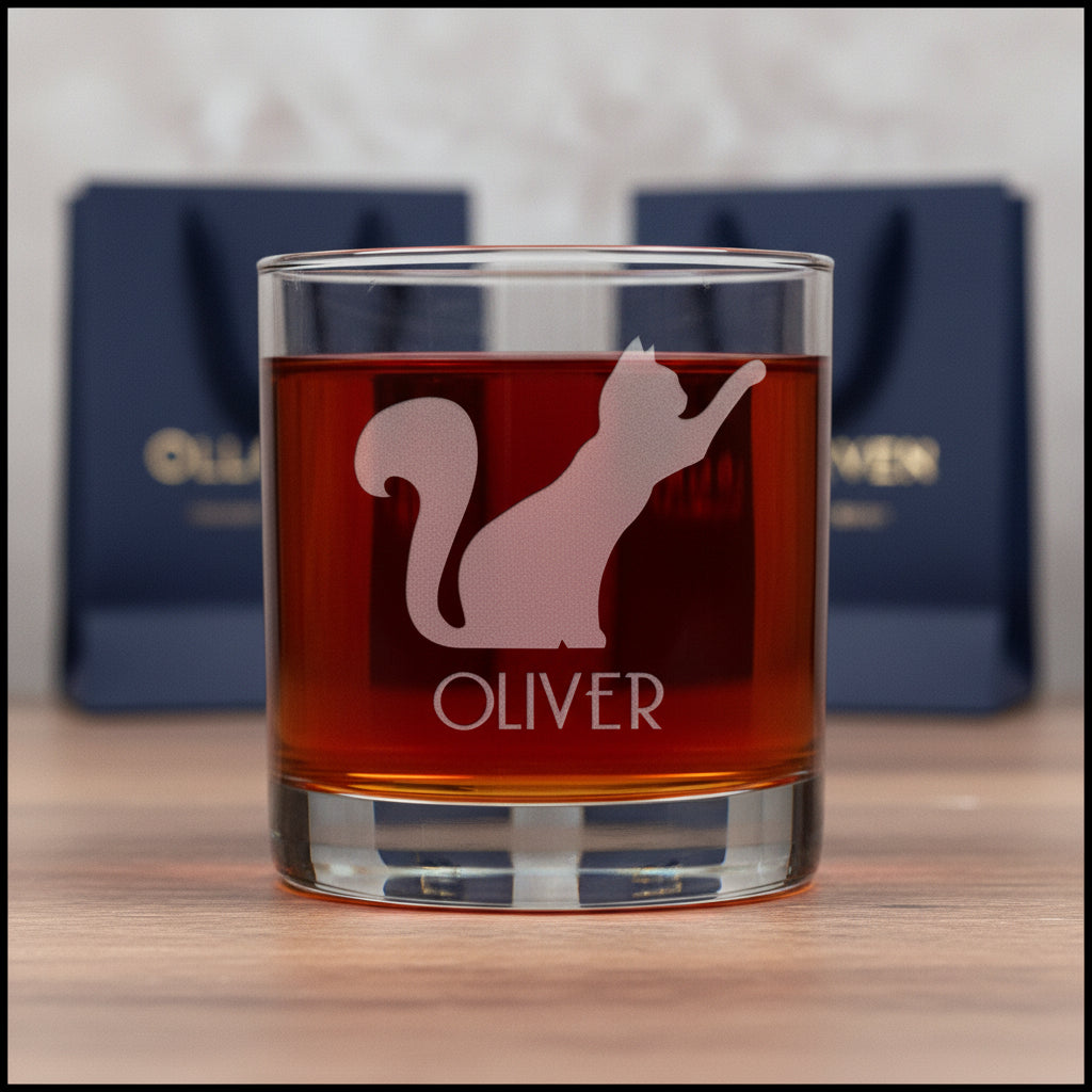 A clear glass rocks tumbler filled with amber liquid, featuring an etched silhouette of a cat and the name OLIVER on the front, set against a textured gray background.