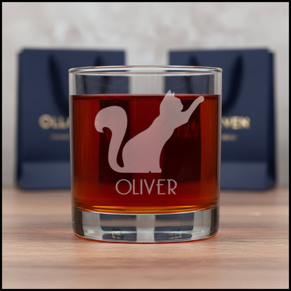 A clear glass rocks tumbler filled with amber liquid, featuring an etched silhouette of a cat and the name OLIVER on the front, set against a textured gray background.
