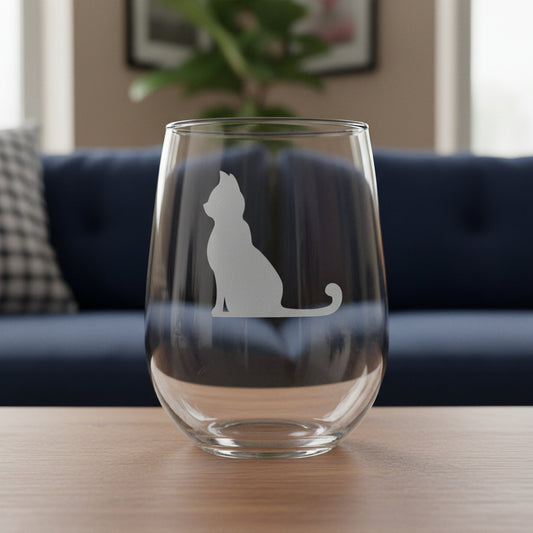 A clear stemless wine glass with a cat silhouette etched on the front, displayed on a wooden surface, with a blue couch in the background.
