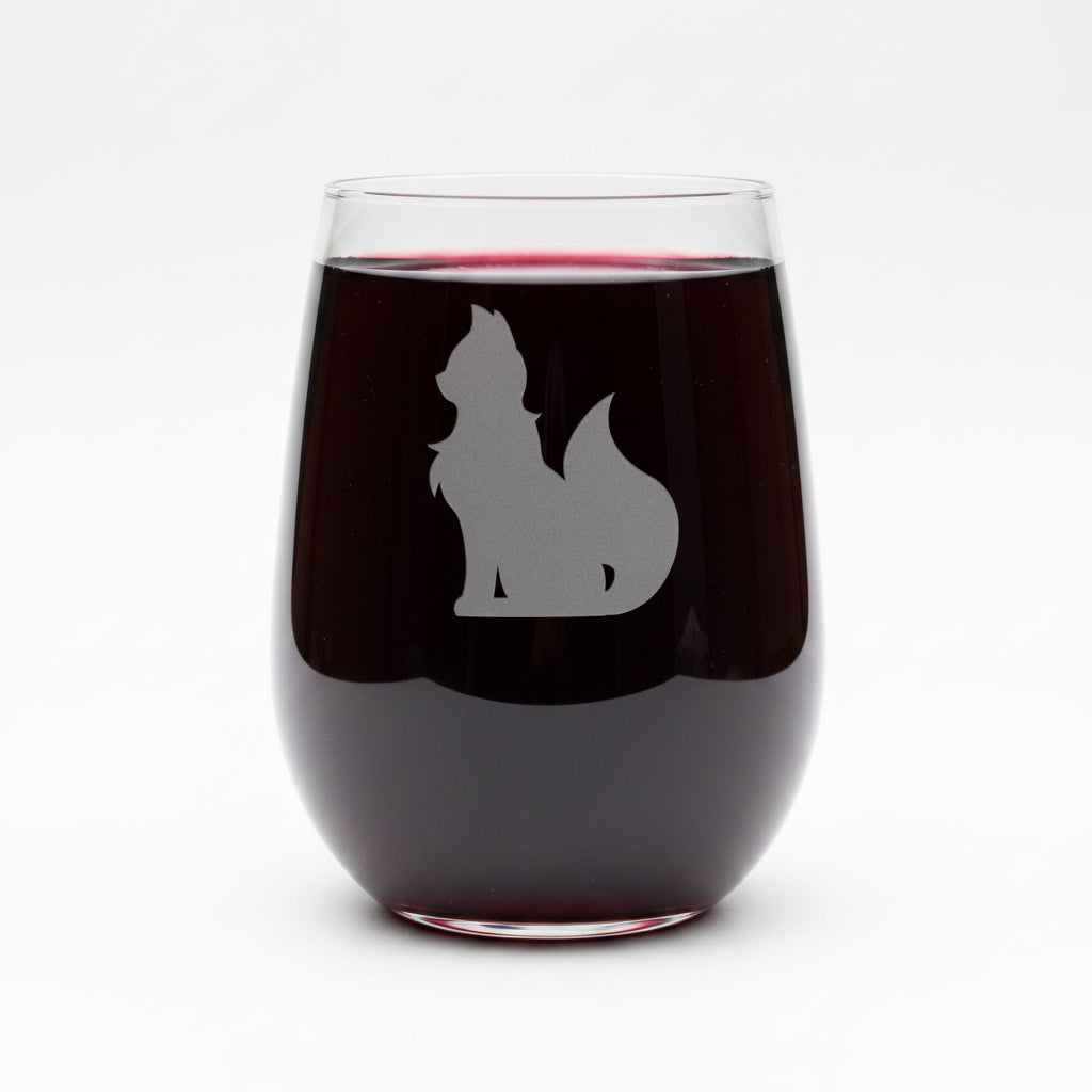 A clear stemless wine glass with an etched silhouette of a cat, shown on a wooden surface against a solid white background.