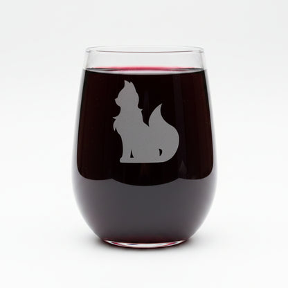 A clear stemless wine glass with an etched silhouette of a cat, shown on a wooden surface against a solid white background.