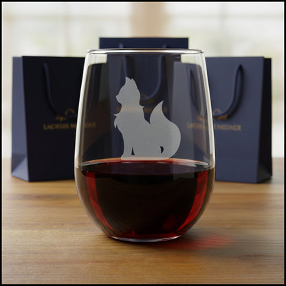 A clear stemless wine glass with an etched silhouette of a cat, shown on a wooden surface with gift bags in the background.