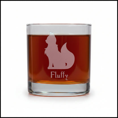 A clear rocks glass filled with an amber-red drink, featuring  an etched cat silhouette of a fluffy dog and the name 'Fluffy' printed below. The glass is centered on a wooden surface against a white  background.