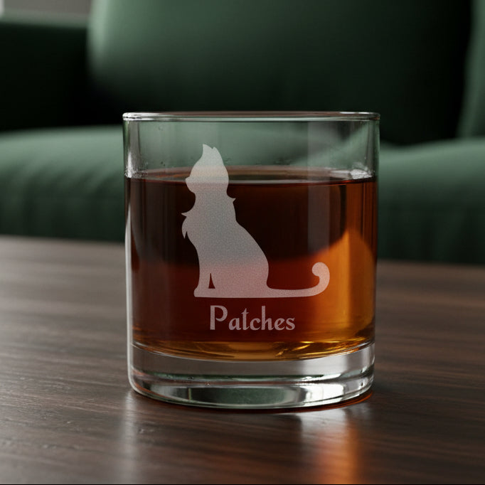 A clear rocks glass filled with an amber-red liquid, featuring an etched cat silhouette and the name 'Patches' on the front, set against a green couch background.