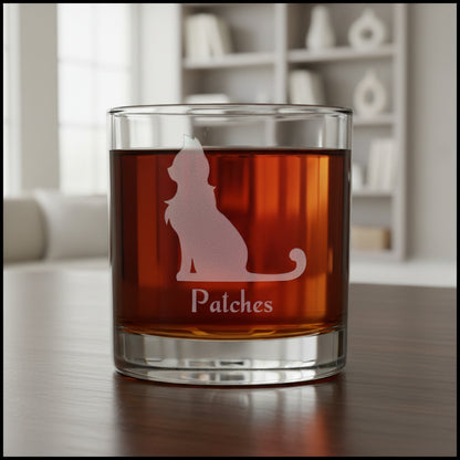 A clear rocks glass filled with an amber-red liquid, featuring a frosted pinkish cat silhouette and the name 'Patches' on the front, set against a textured gray background.