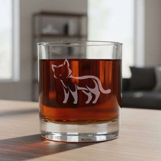 A clear rocks glass filled with an amber-red drink, featuring an etched cat silhouette on the front, set against a living room background.