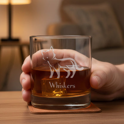 A hand holdin a clear rocks glass with an etched cat silhouette and the name 'Whiskers' on the front, filled with an amber-red drink.